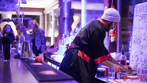 A bartender at a bar with a chalkboard menu, bottles, and glasses on the counter. - Marco Polo Malta Hostel (St. Julian's)