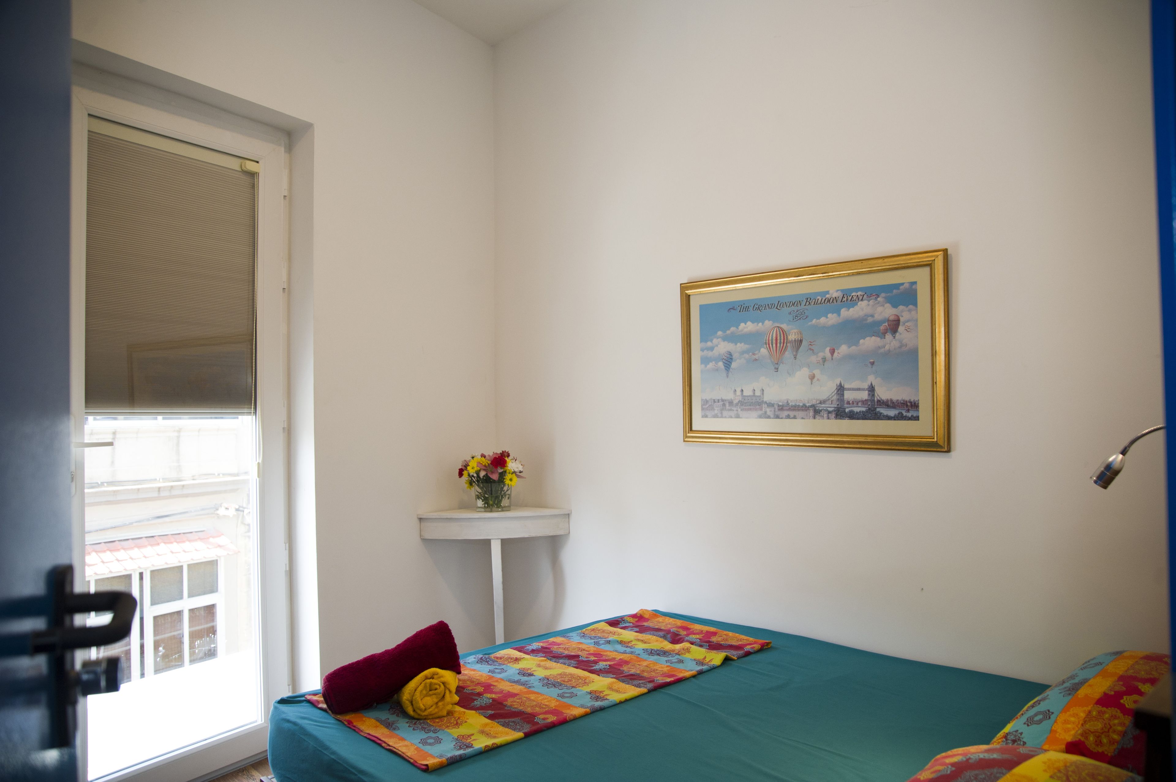 A small room with a bed, a window, a framed picture on the wall, and a small table with flowers.