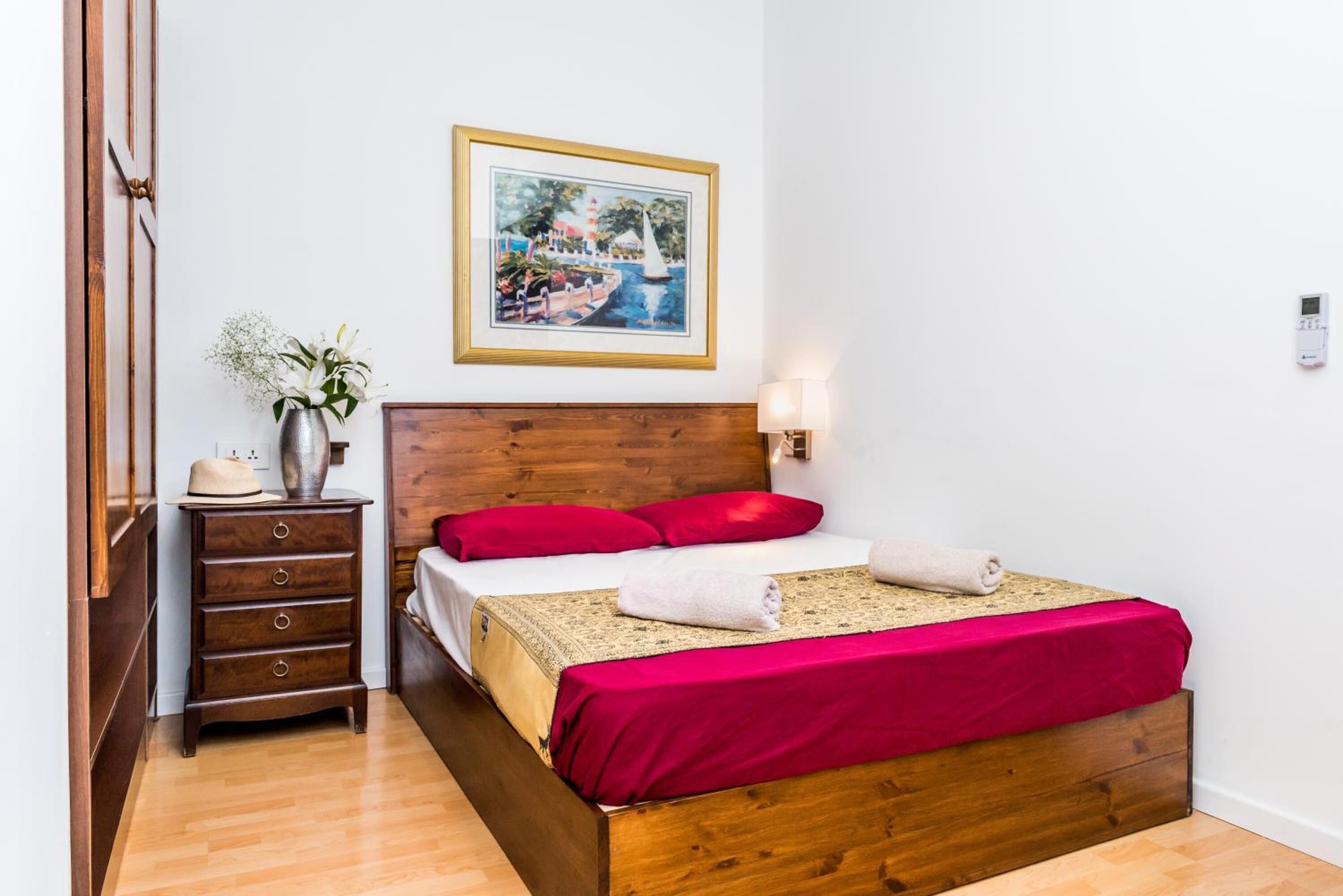 Una camera da letto ordinata con un letto in legno, una piccola cassettiera, un vaso con fiori, un quadro incorniciato e un termostato a parete.
