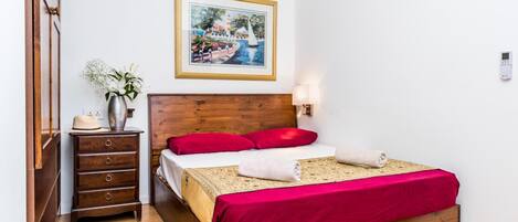 A neatly arranged bedroom with a wooden bed, a small dresser, a vase with flowers, a framed painting, and a wall-mounted thermostat.