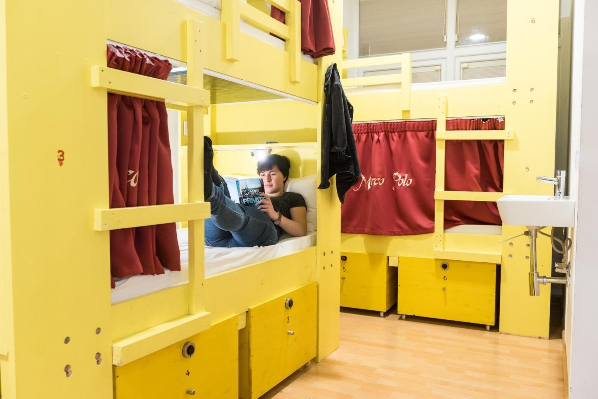 A bunk bed room with a person lying on the lower bunk. There are red curtains on the windows, and a sink is visible to the right.