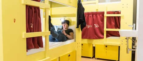 A bunk bed room with a person lying on the lower bunk. There are red curtains on the windows, and a sink is visible to the right.