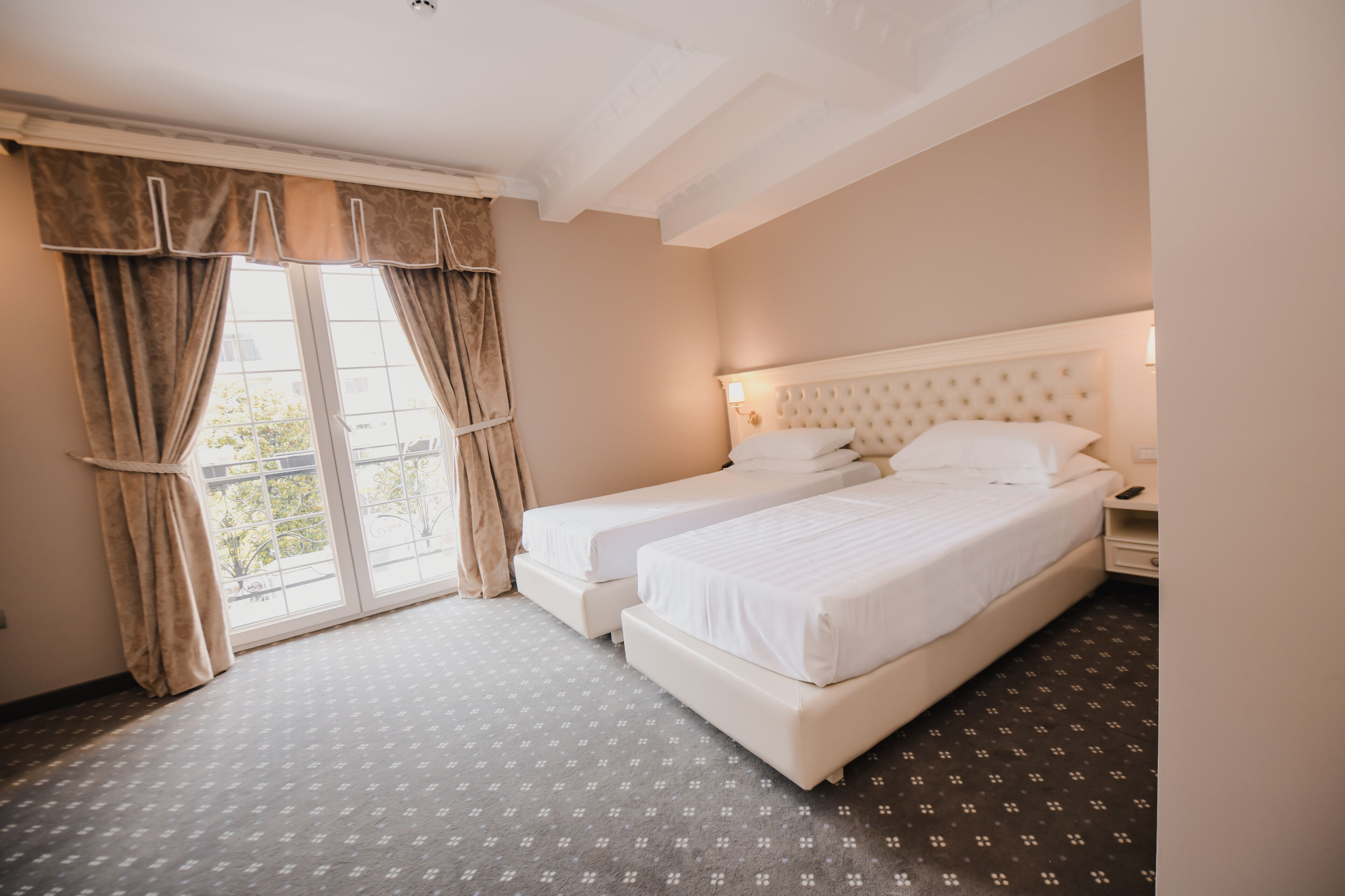 A hotel room with two beds, a large window with curtains, a white headboard, and a patterned floor.