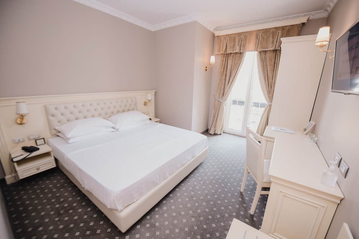 Una habitación de hotel con una cama grande, una mesita de noche con lámpara, un aparador con espejo y una ventana con cortinas.