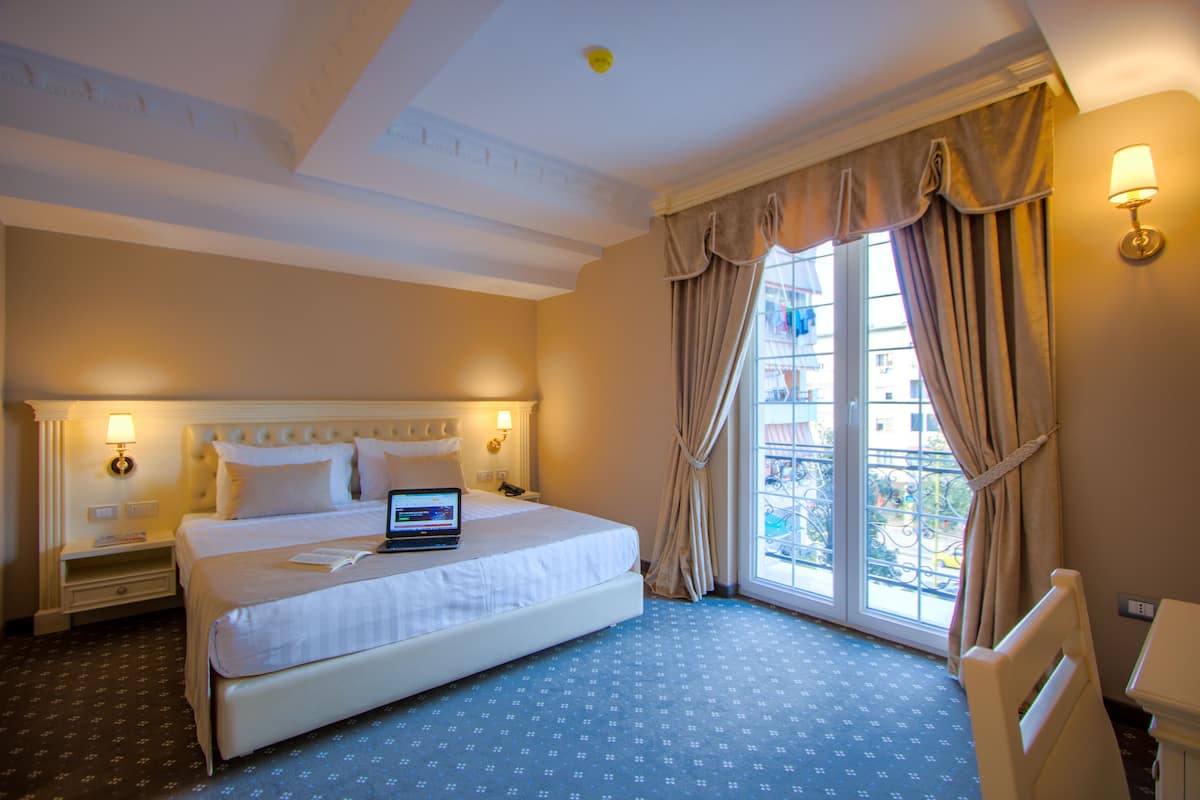 Habitación de hotel con una cama grande, una computadora portátil sobre la cama, una mesita de noche, una silla y una ventana con cortinas.