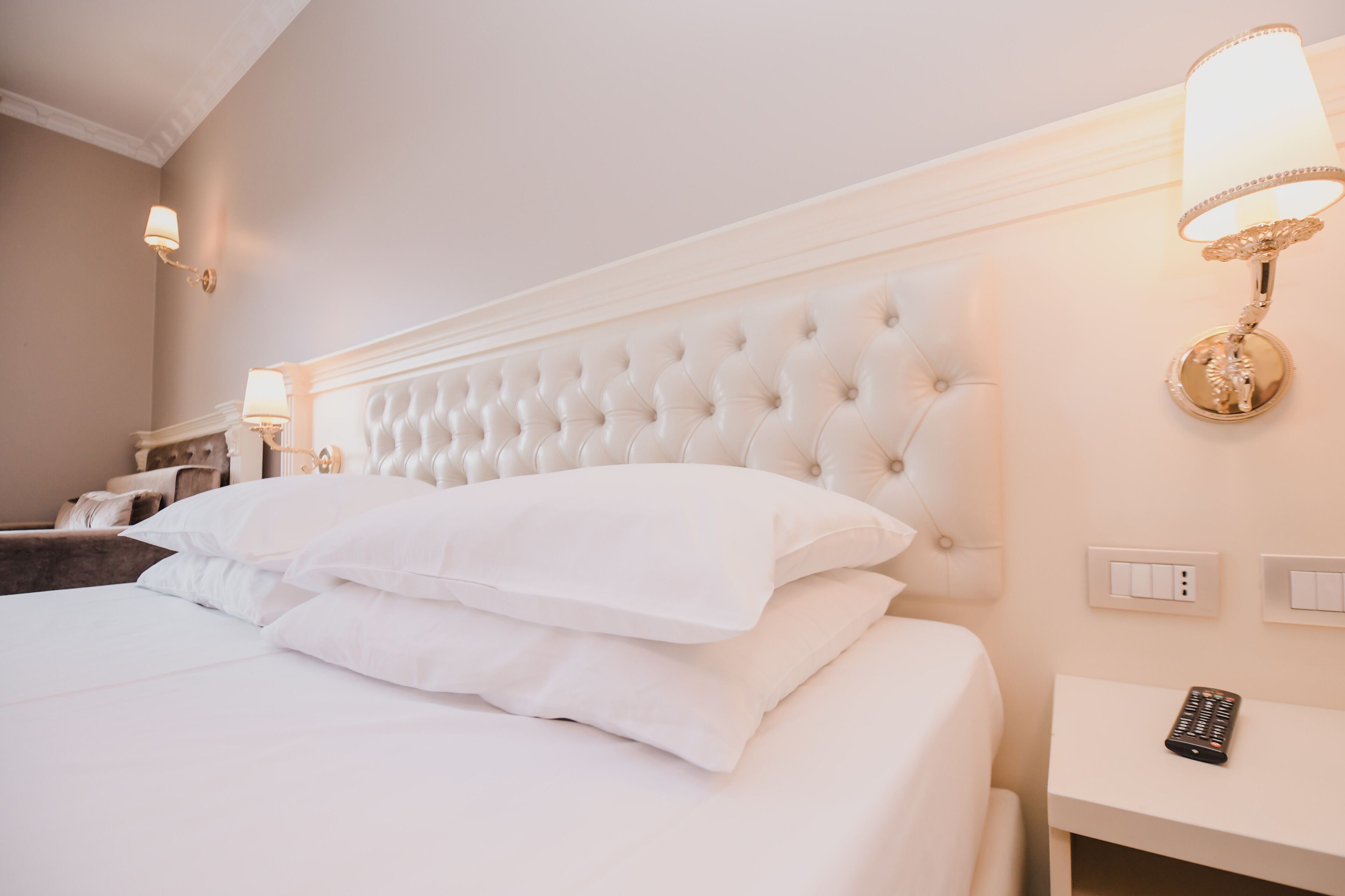 A hotel room with a tufted headboard, white bedding, a bedside table with a remote control, and wall-mounted lamps.