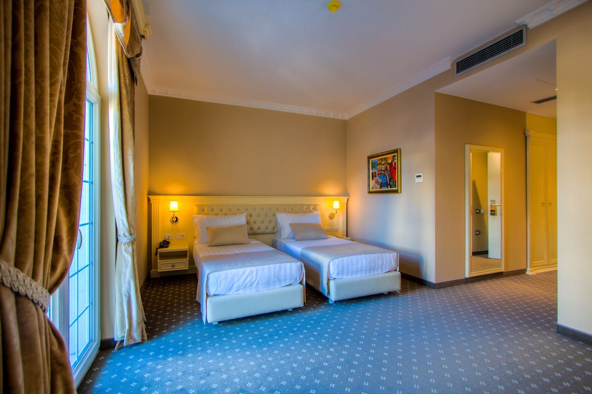 Habitación de hotel con dos camas individuales, una mesita de noche, una lámpara fijada a la pared y una puerta que da a otra habitación.
