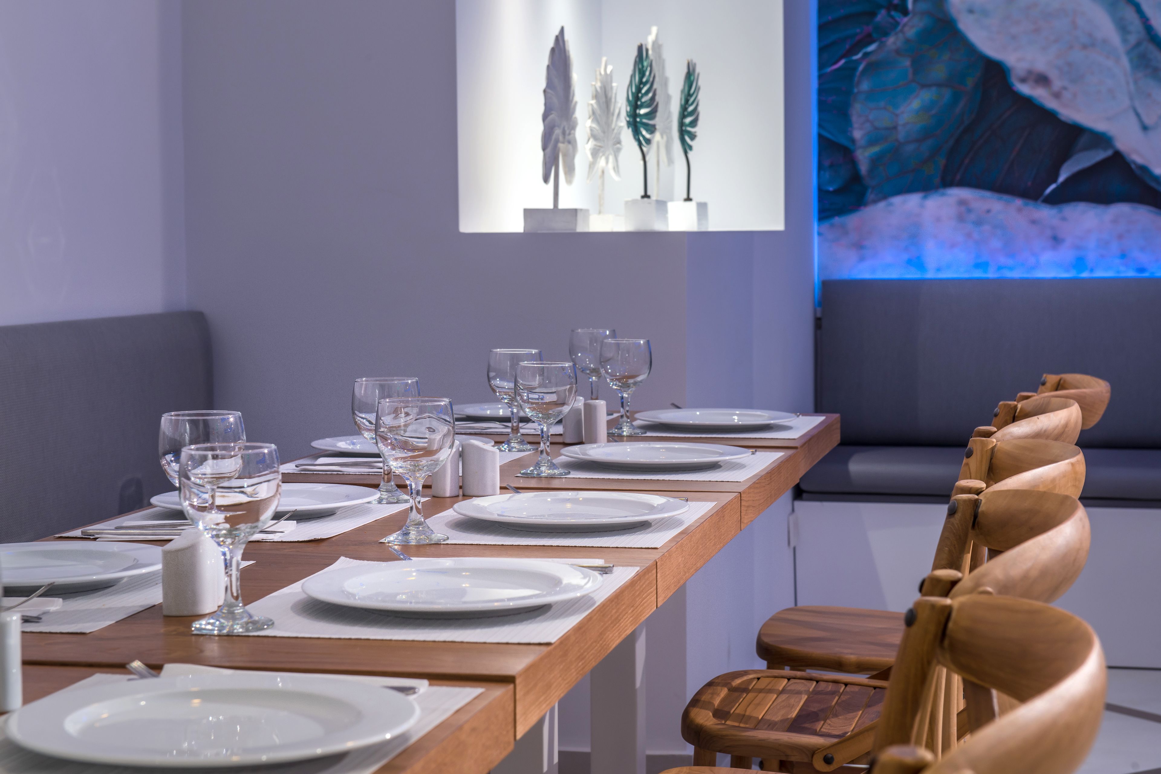 A dining table set with plates, glasses, and cutlery, with a wooden chair and a wall-mounted artwork in the background.