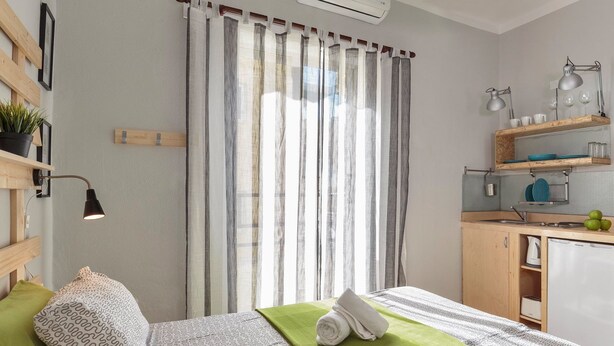 Una habitación de hotel moderna con una cama bien hecha, un plaid verde, una toalla y una pequeña cocina con un lavamanos y un hervidor.