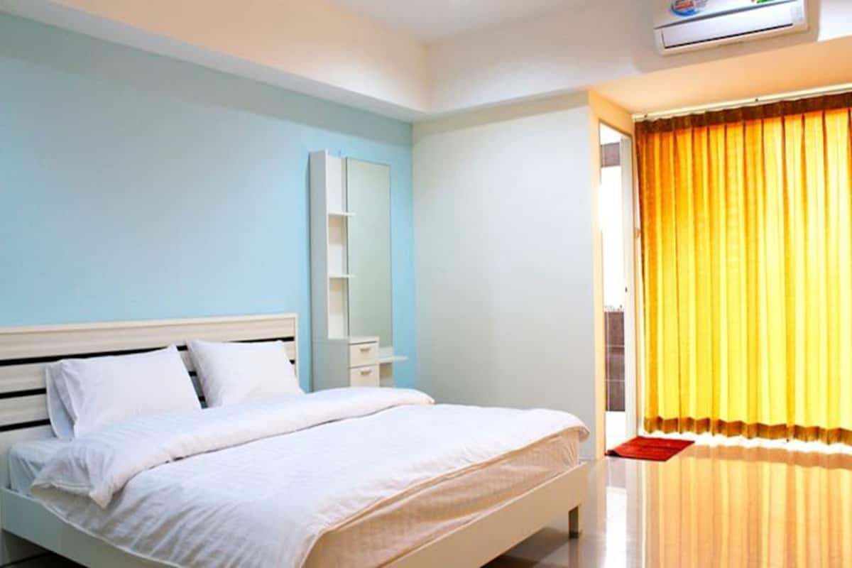 Un dormitorio con cama, mesita de noche, espejo y ventana con cortinas amarillas.