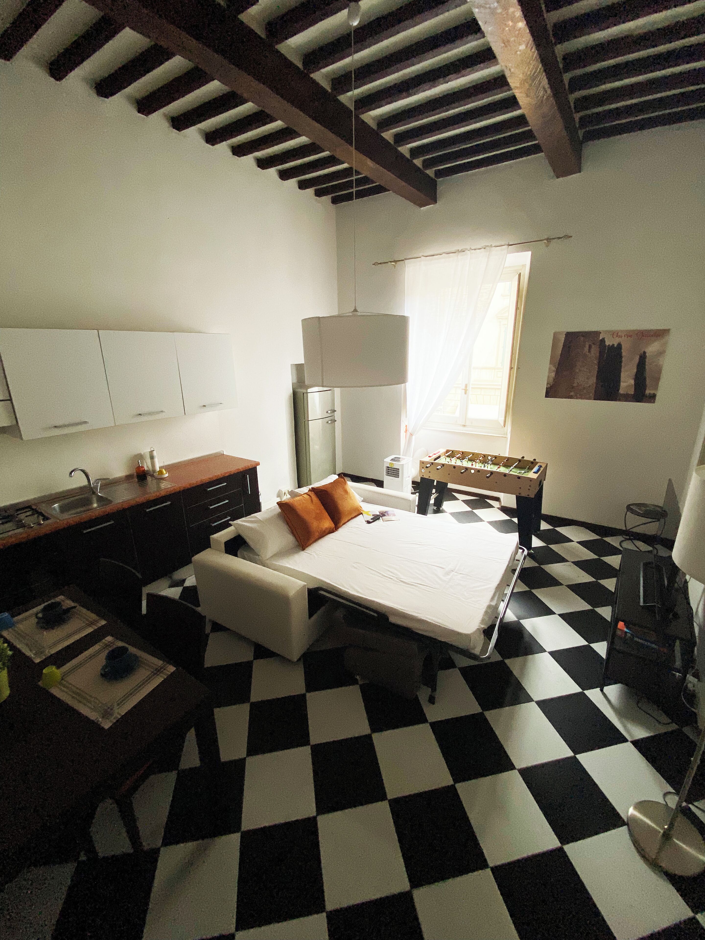 Habitació d'hotel amb un sòl de pissos, un llit amb matalàs blanc, una taula de nit amb un ordinador, un frigorífic i una finestra amb cortines.