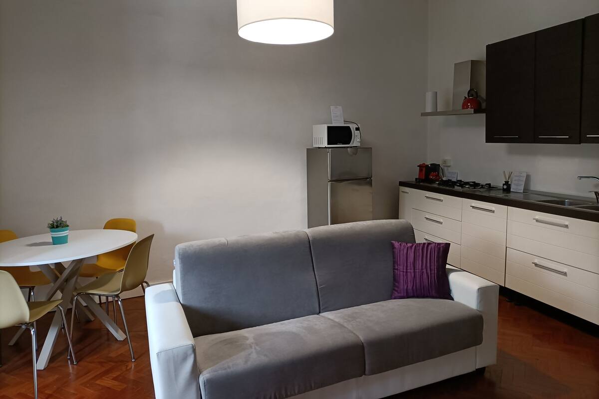 Una cocina moderna con un sofá gris, una mesa de comedor con sillas amarillas y una lámpara colgante blanca.