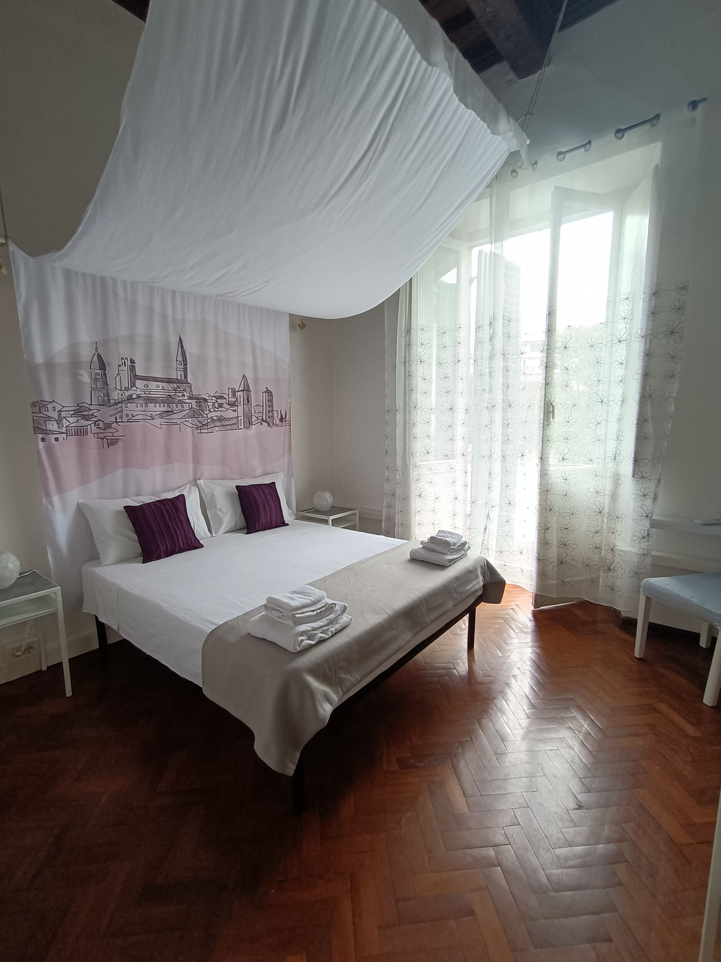 Un dormitorio con una cama grande, ropa de cama blanca, dosel, mesita de noche, una silla y una ventana con cortinas florales.