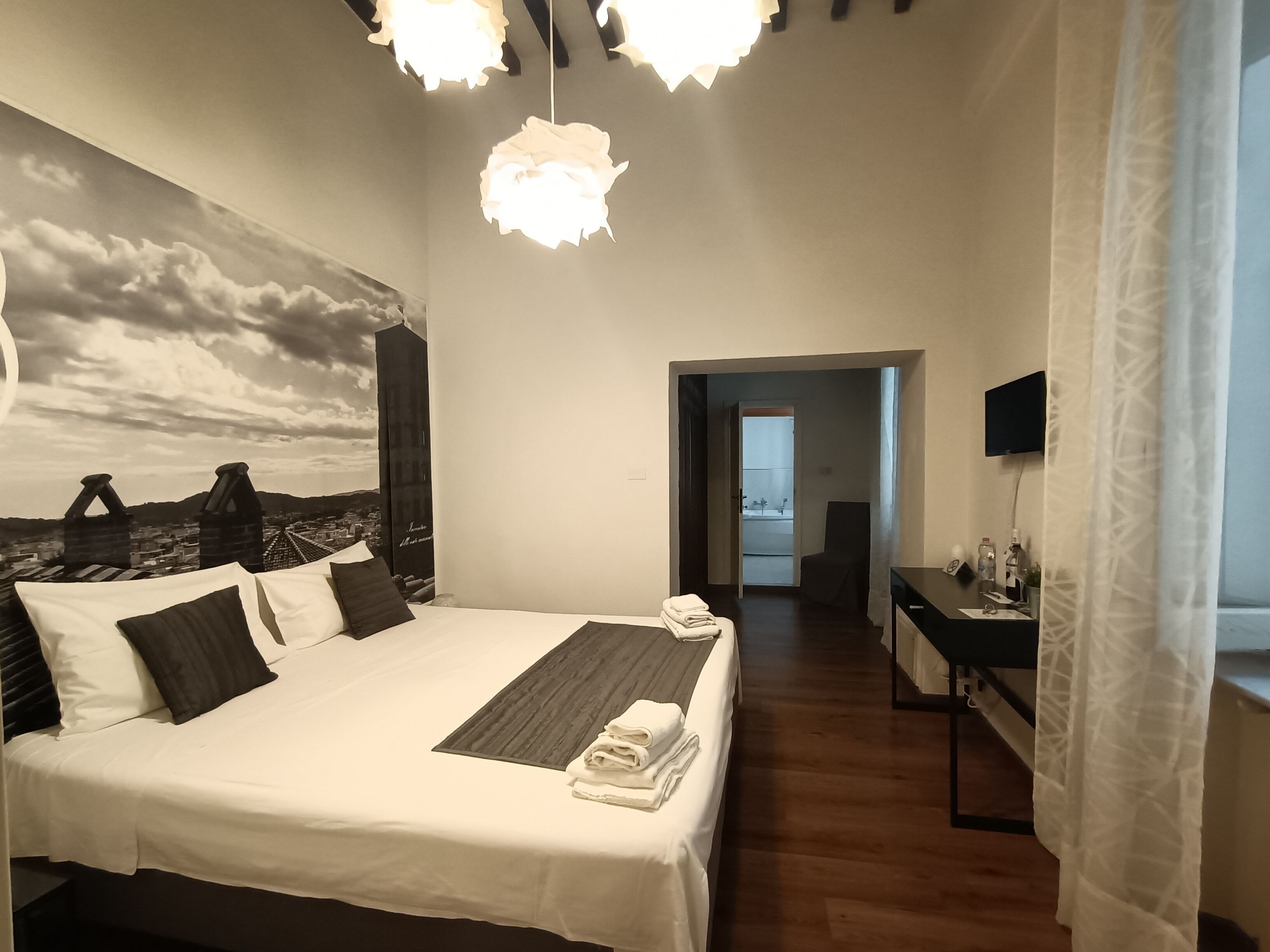 Een slaapkamer met een groot bed, uitzicht op een stadsbeeld, een bureau met een computer en een televisie aan de muur.