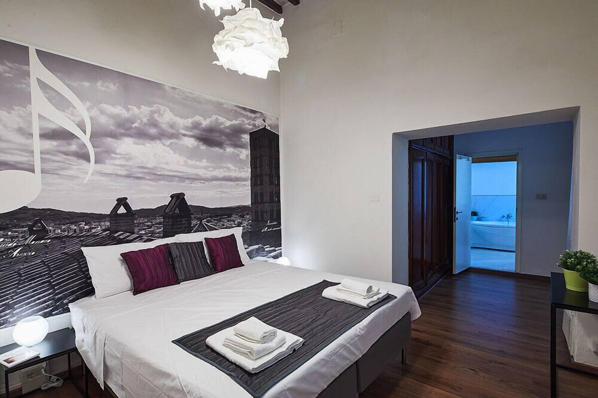 Habitación de hotel moderna con una cama grande, un mural de paisaje urbano en la pared y vista de la ciudad a través de una puerta abierta.
