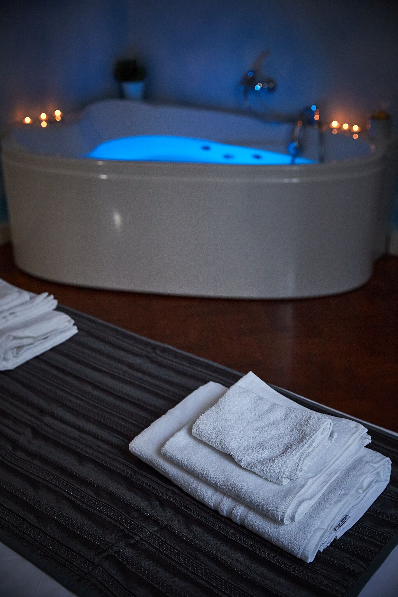 A bathtub with blue lighting, a wooden bench with folded towels, and a dark wooden floor.