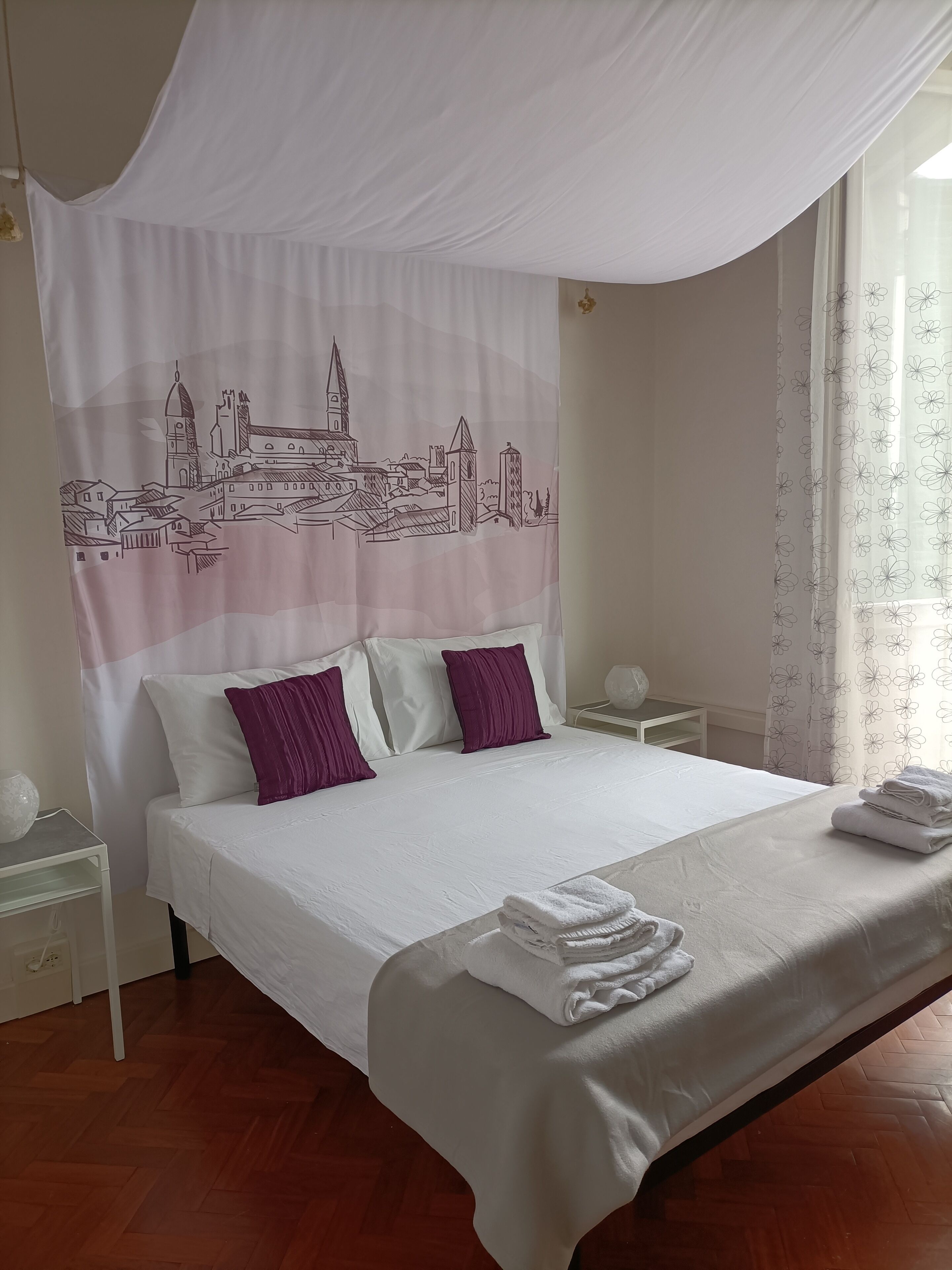 Ein ordentlich bezogenes Bett mit weißen und violetten Kissen, einem weißen Baldachin und Blick auf eine Stadtlandschaft durch den Baldachin.