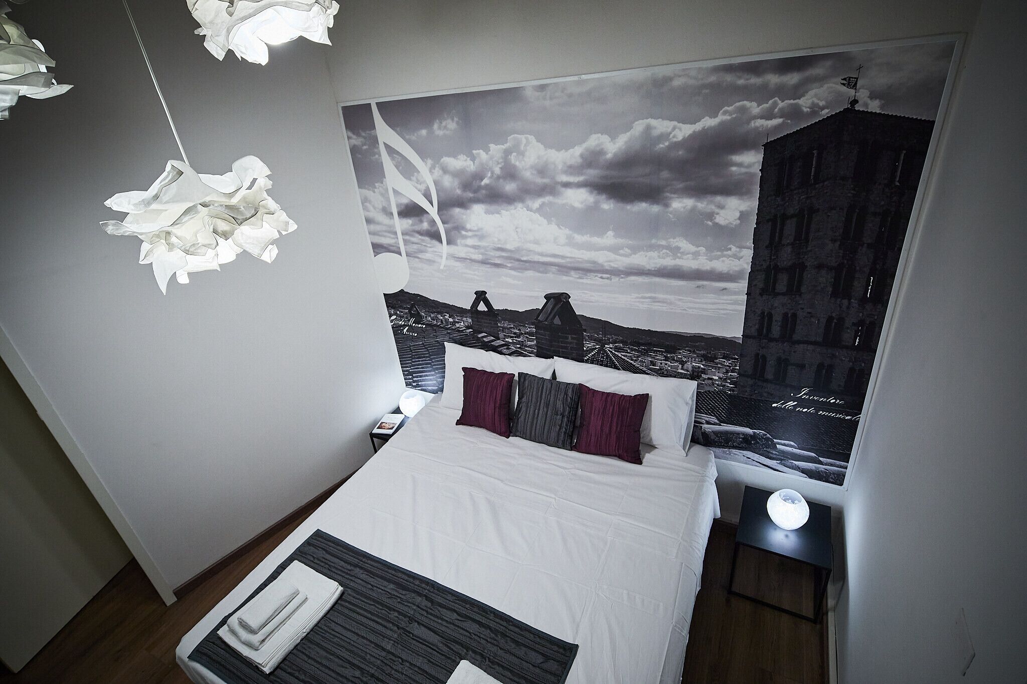 A modern hotel room with a large bed, a nightstand, a bedside table with a lamp, and a wall-mounted artwork depicting a cityscape.
