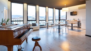 Un vaste espace de vie comprenant un magnifique piano à queue, un canapé confortable et un coin repas avec plusieurs chaises et tables. De grandes fenêtres offrent une vue sur le paysage urbain.