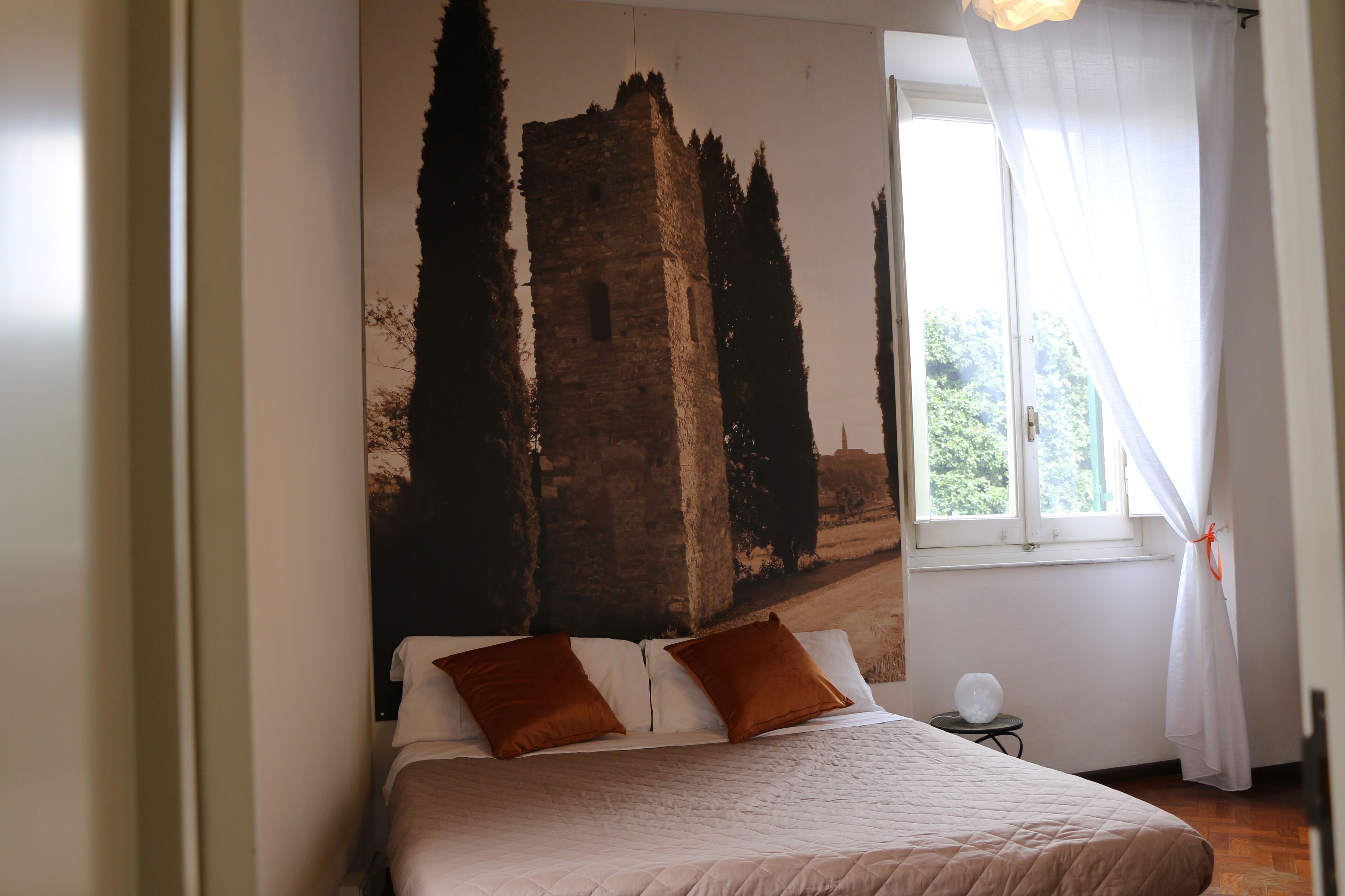 Un dormitorio con una cama, dos almohadas, una mesita de noche, una ventana con cortinas y un mural en la pared con un castillo.
