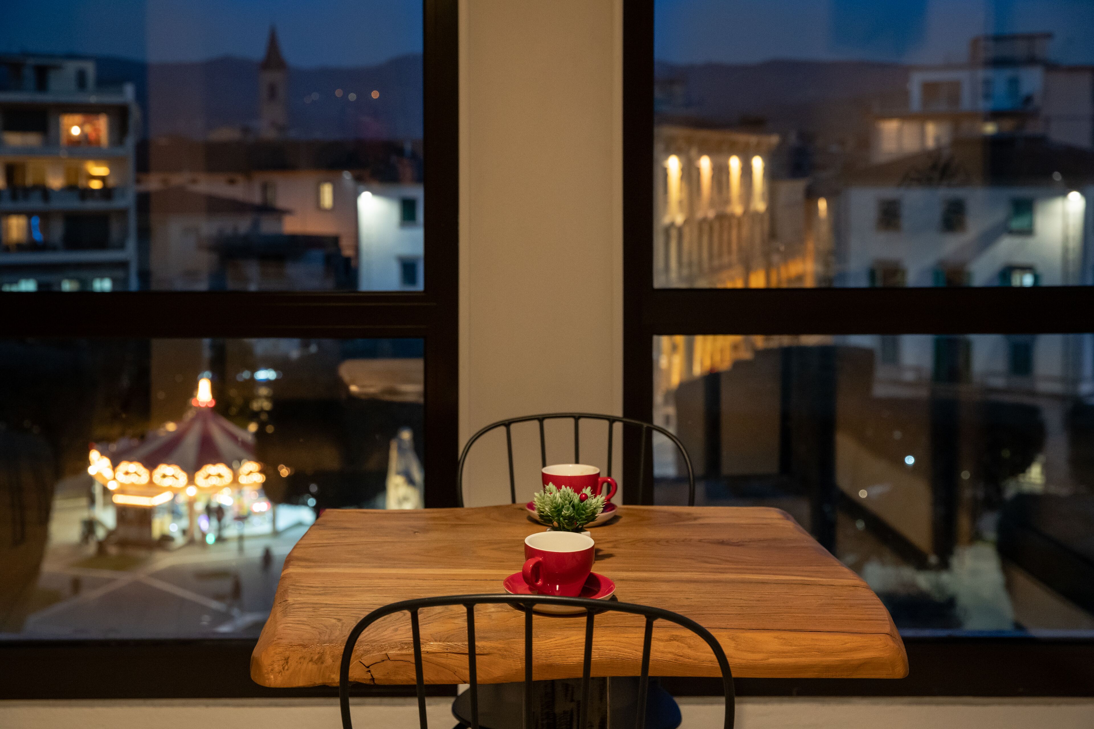 A wooden table with two red cups and a small plant, set against a window overlooking a cityscape with buildings and a distant carousel.