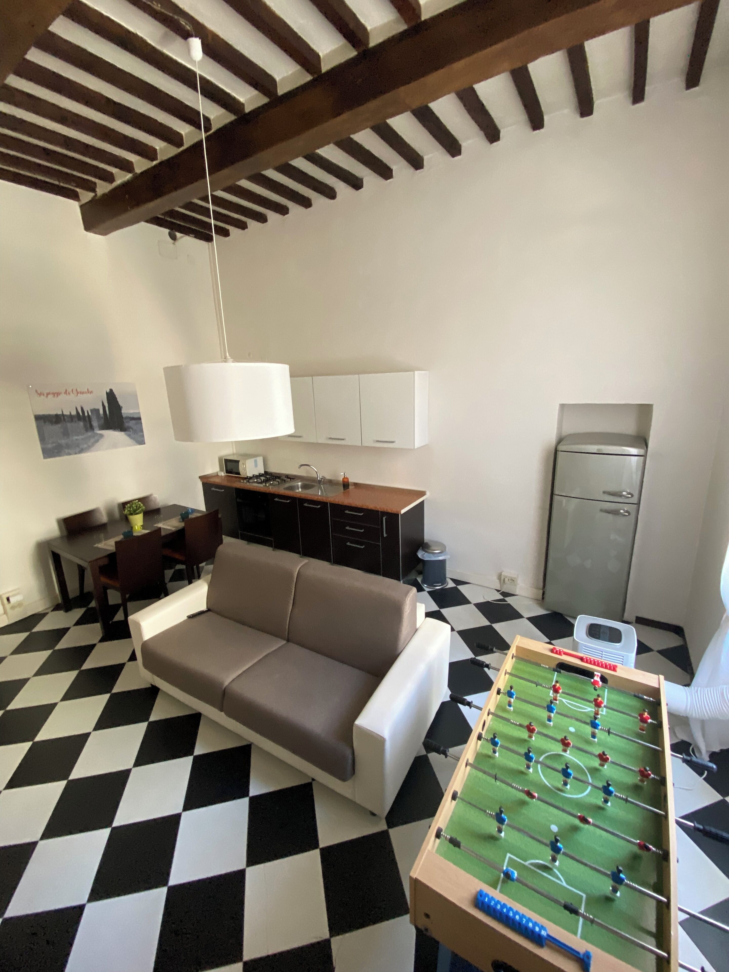 A living room with a sofa, a foosball table, a dining table with chairs, a kitchen area with cabinets, a refrigerator, and a ceiling with exposed beams.