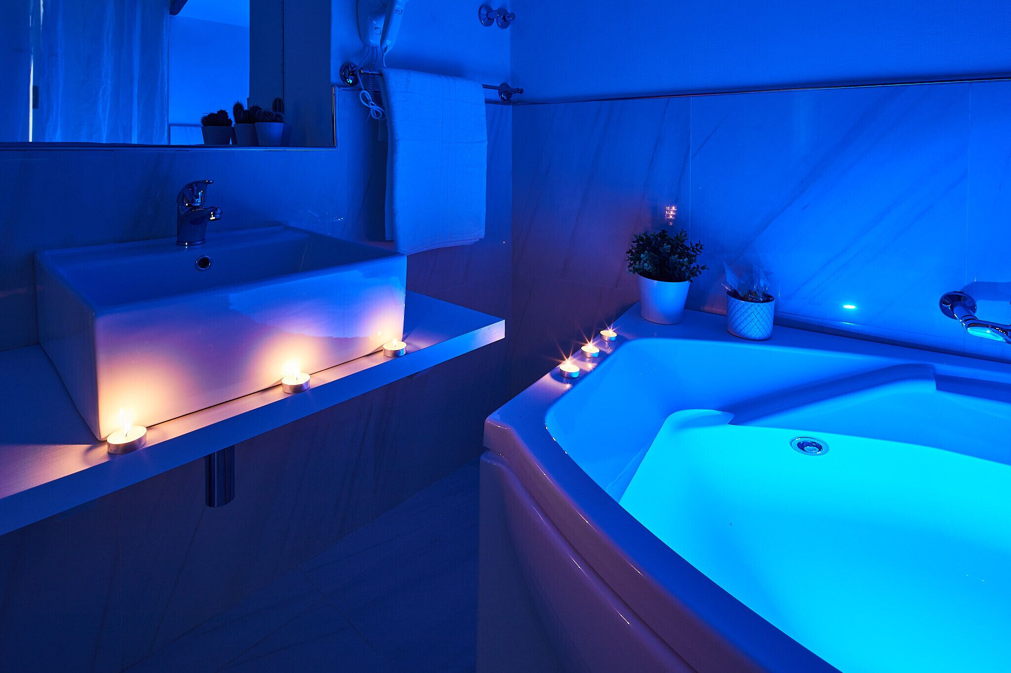 A bathroom with a large bathtub and a sink, both illuminated by blue lighting. There are candles placed on the sink and along the edge of the bathtub.
