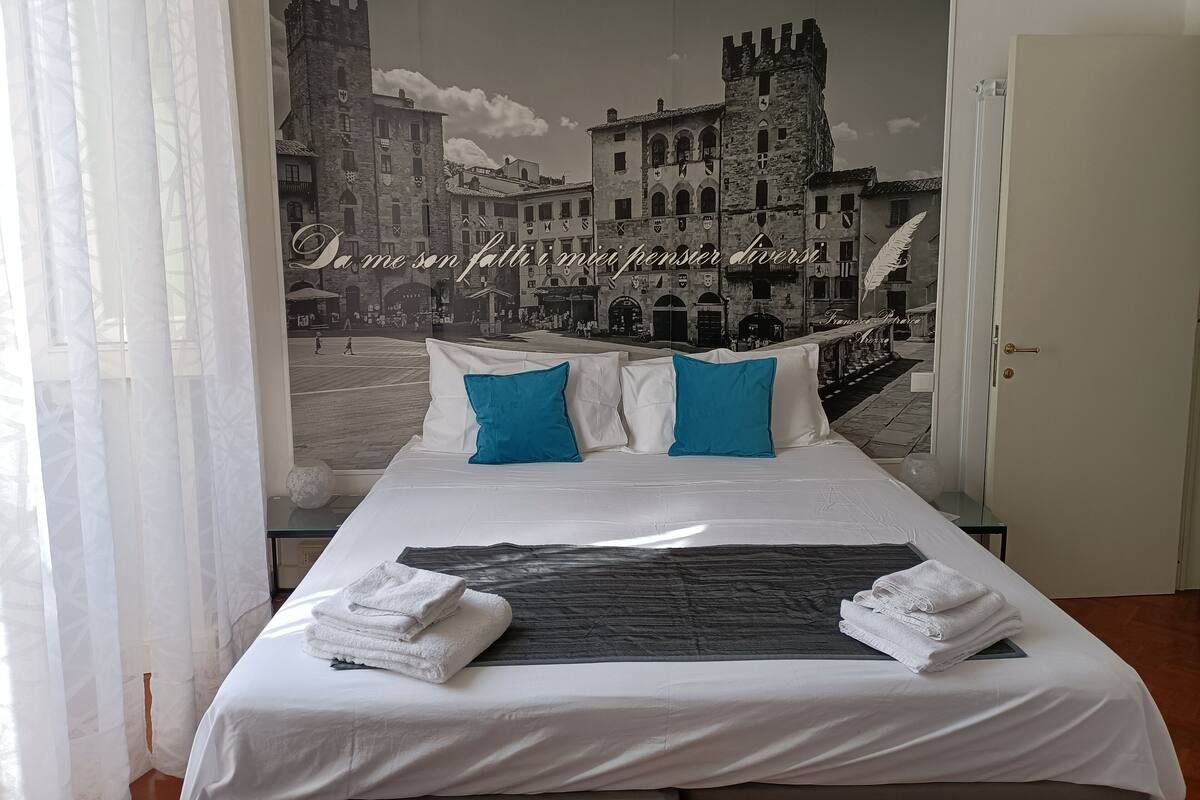 Habitación de hotel con una cama grande, dos almohadas azules y vista a un pueblo histórico a través de un ventanal.