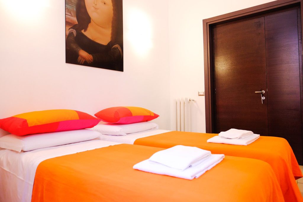 Een hotelkamer met twee bedden, elk voorzien van oranje beddengoed en witte handdoeken. Er is een houten deur en een radiator aan de muur.