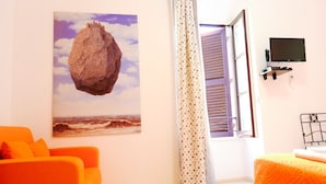 Una habitación con cama, televisor montado en la pared, un cuadro de rocas y olas, y una ventana con persianas.