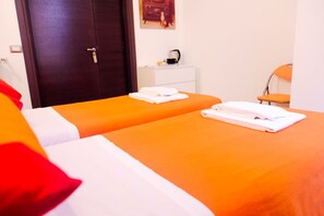 A hotel room with two beds, each with an orange bedspread and white linens. There is a white nightstand with a coffee maker, a chair, and a door with a handle. - Your Rome (Rome)