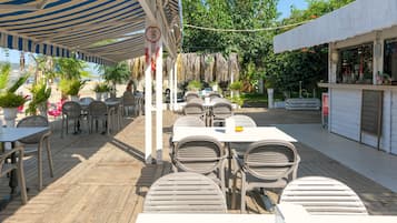 An outdoor patio area with tables and chairs under striped canopies, surrounded by potted plants and a view of the beach.