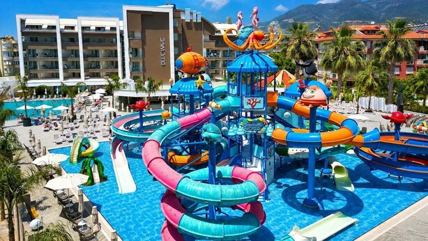Een waterpark met glijbanen en speelconstructies, omringd door een zwembadgedeelte met ligbedden en parasols.