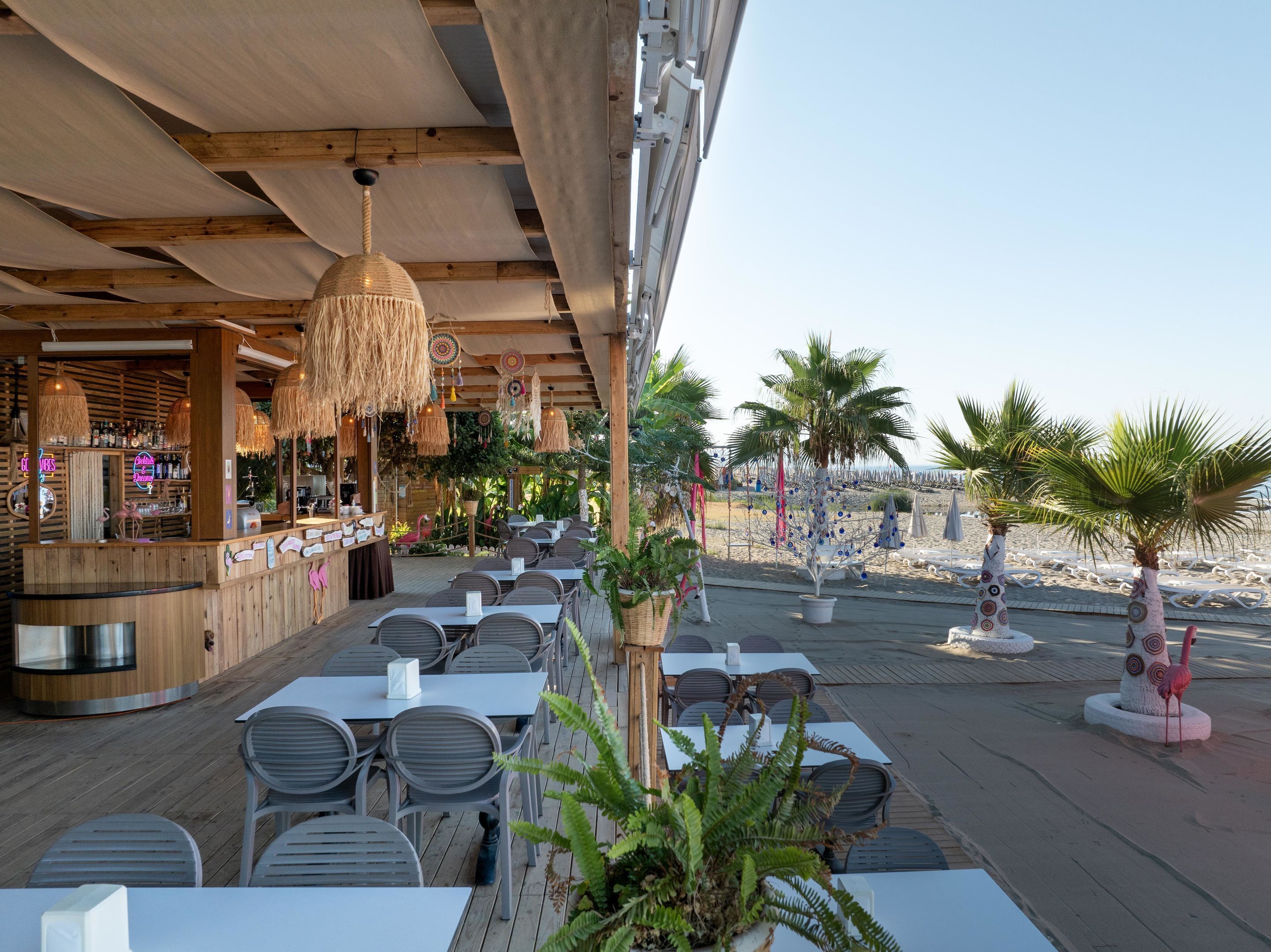 An outdoor dining area with wooden tables and chairs, a bar with a neon sign, and palm trees.