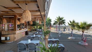 An outdoor dining area with wooden tables and chairs, a bar with a neon sign, and palm trees.
