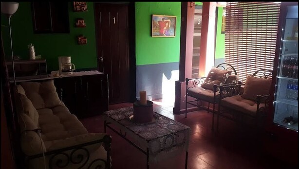 Una sala de estar con un sofá, una mesa de centro y un comedor con una mesa y sillas. Hay un cuadro en la pared y una puerta con un número.