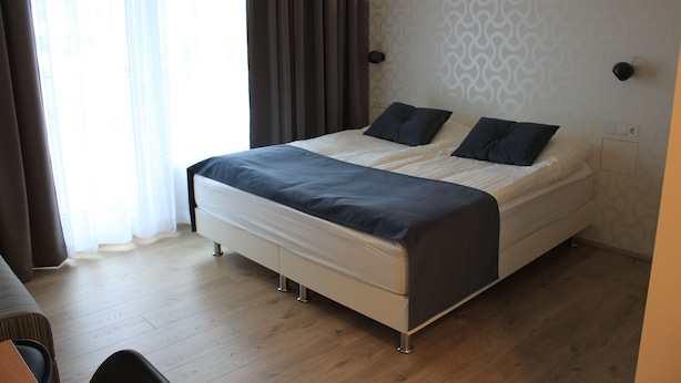 Una habitación de hotel moderna con una cama grande, una mesita de noche y un ventanal con cortinas.