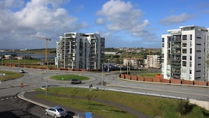 Un moderno edificio de apartamentos con varios pisos y balcones, ubicado cerca de una glorieta con una carretera y una grúa de construcción al fondo.