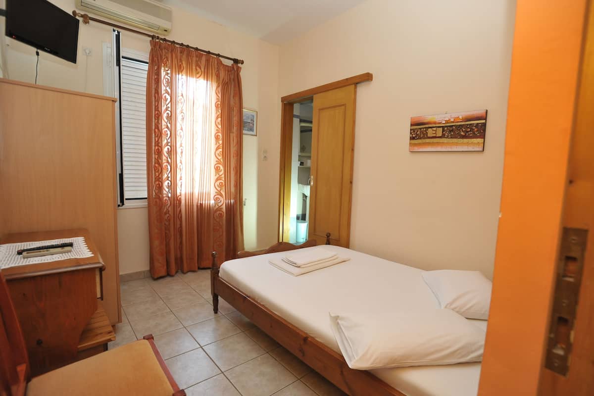 A small, neatly arranged bedroom with a single bed, a wooden desk, a chair, a wardrobe, and a television mounted on the wall.