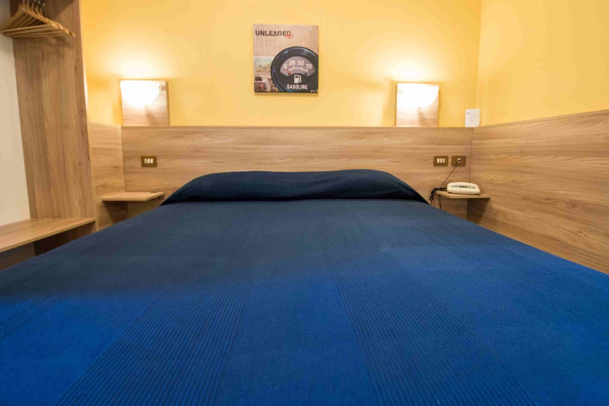 A hotel room with a bed, a nightstand, a wall clock, and a poster on the wall.