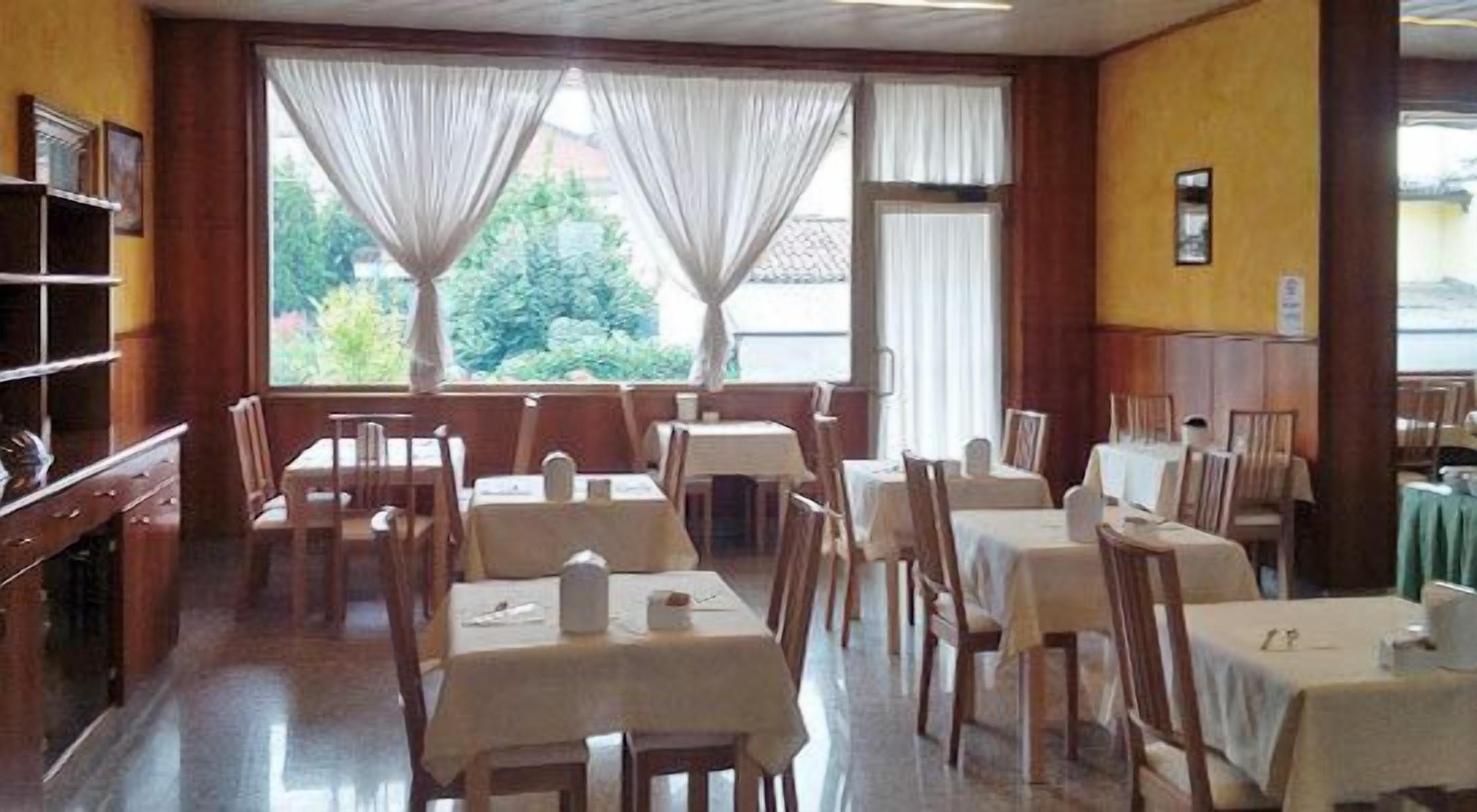 Un restaurante con varias mesas y sillas, amplias ventanas con cortinas y vistas a zonas verdes.