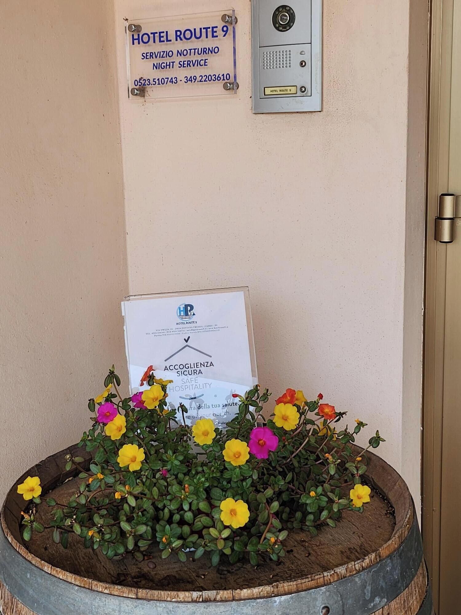 Letrero del Hotel Route 9 con números de contacto, aviso de servicio de seguridad y un arreglo floral sobre un barril de madera.