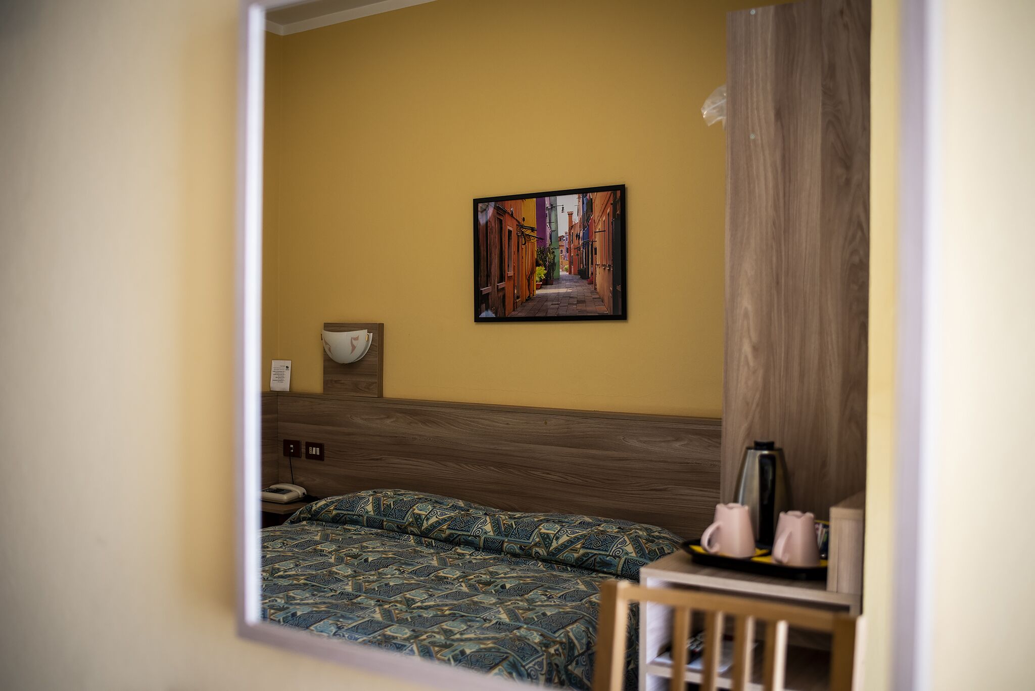 Una habitación de hotel con una cama, una mesita de noche con una lámpara, una cafetera y un cuadro en la pared.