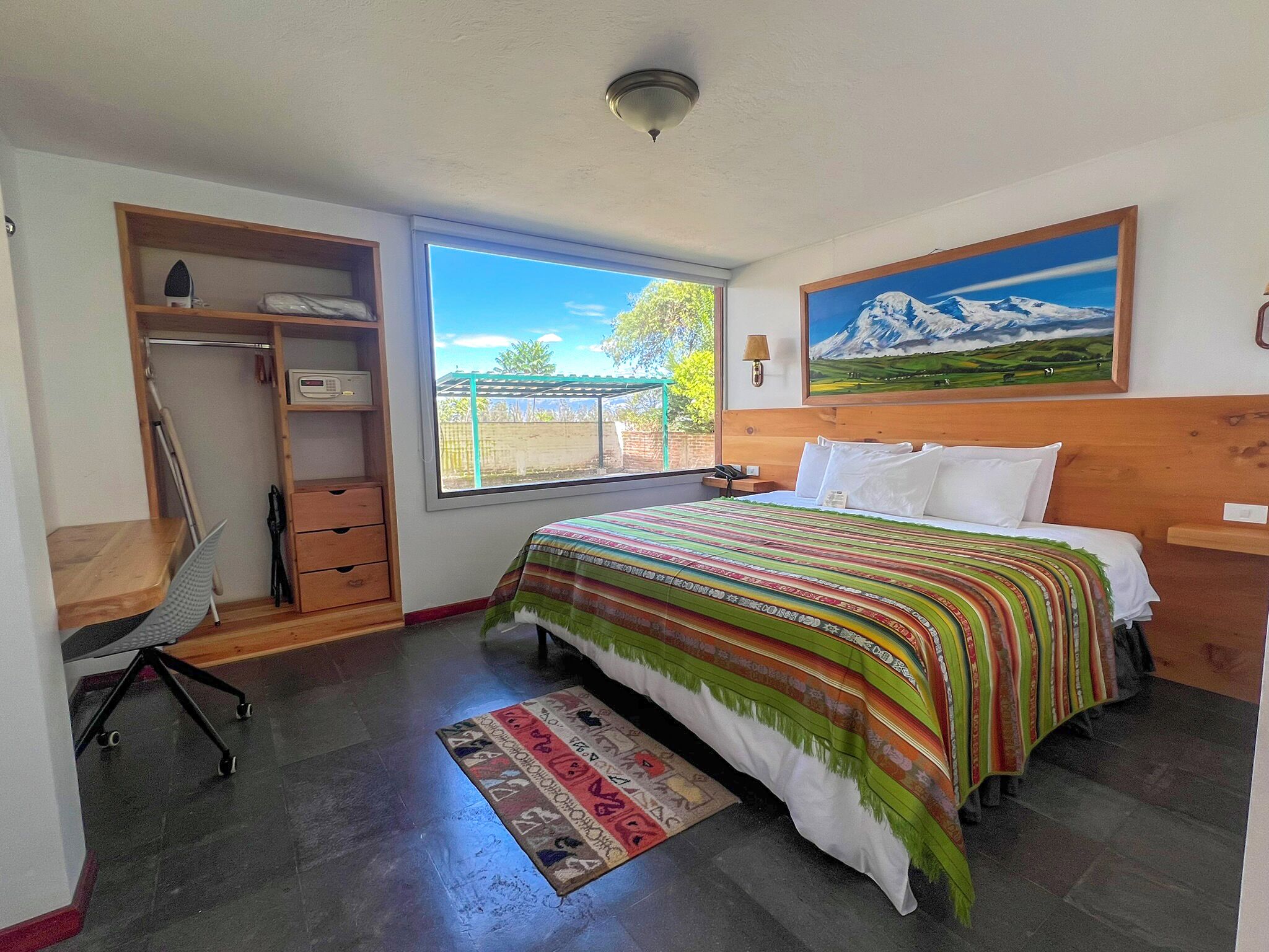 Un dormitorio con cama, escritorio, silla, armario, ventana con vistas y un cuadro en la pared.