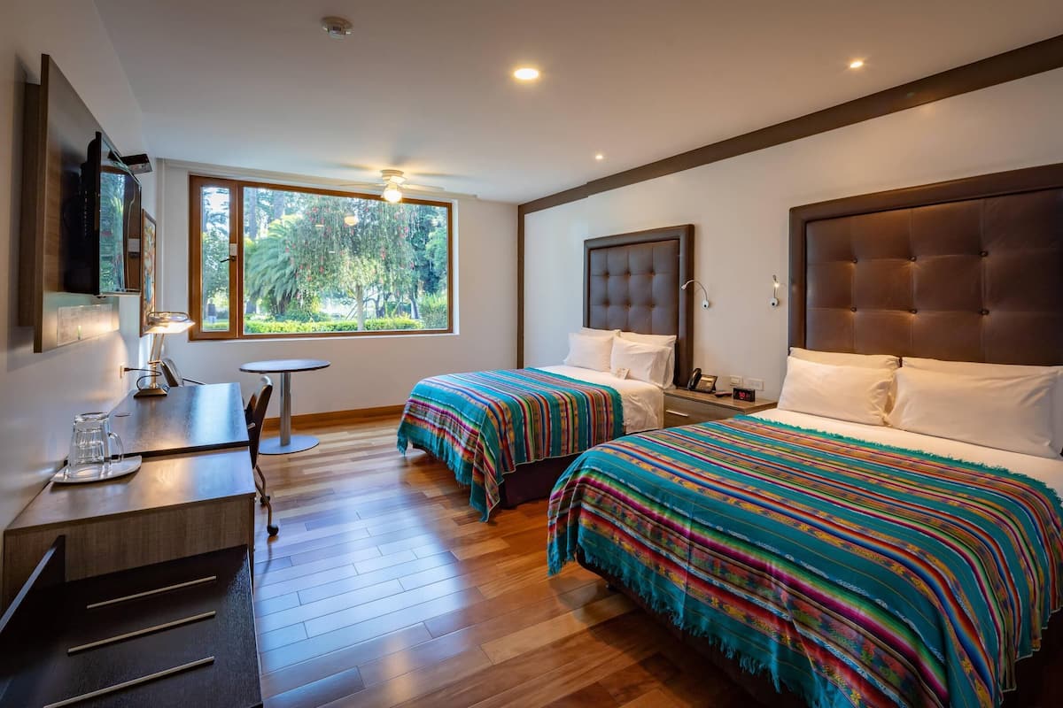 A hotel room with two beds, a desk with a chair, a small table, and a large window offering a view of trees.