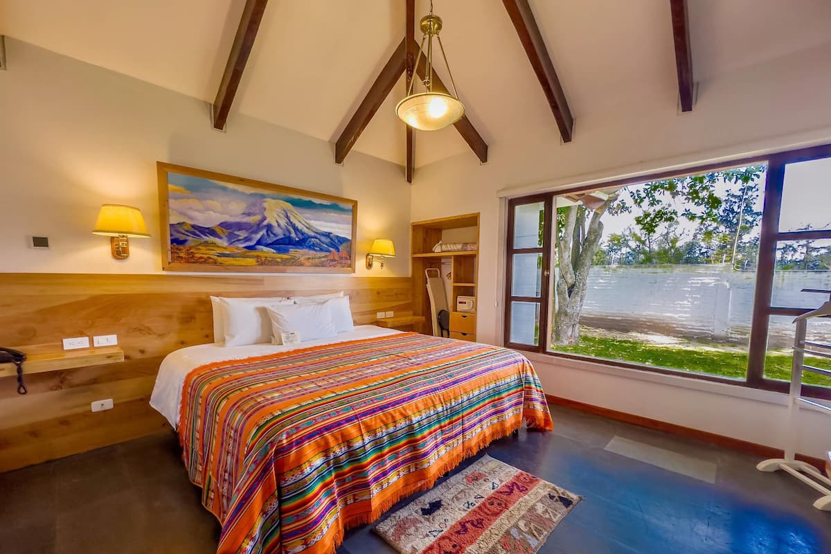 A bedroom with a large bed, a painting of a mountain, a window with a view of a parking lot, and a ceiling fan.