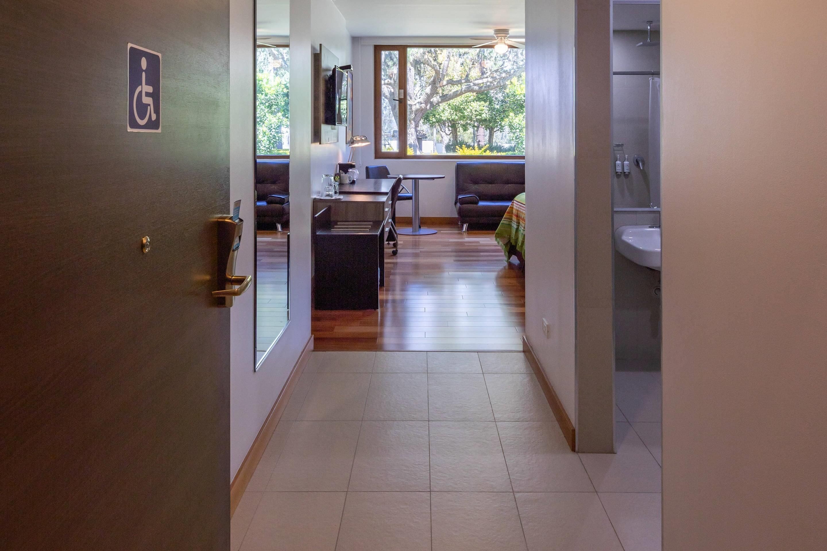 A hallway leading to a dining area with a wheelchair-accessible sign, a dining table, chairs, and a view of trees outside.