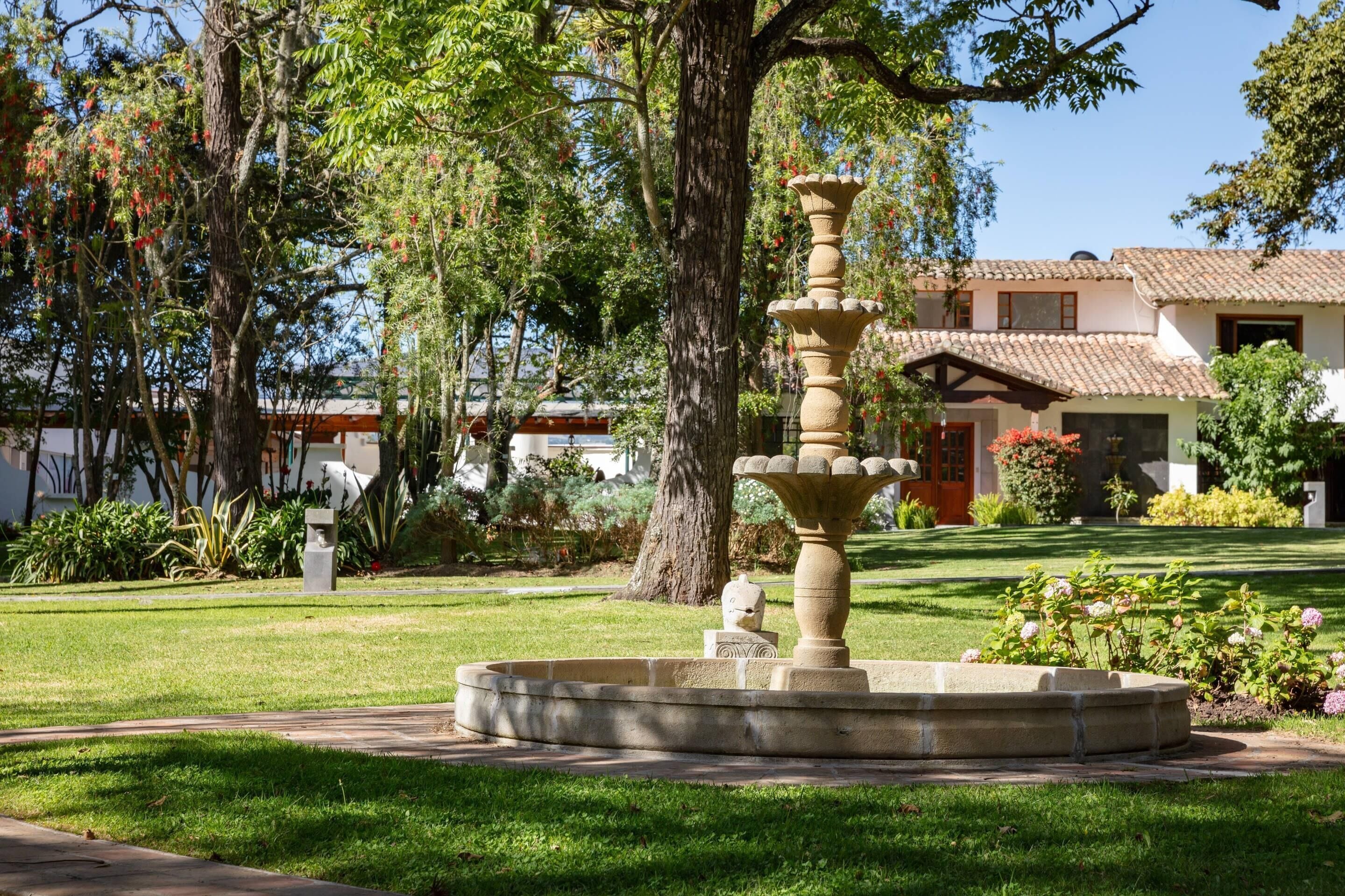 A garden with a stone fountain, a large tree, and a building with a tiled roof in the background.