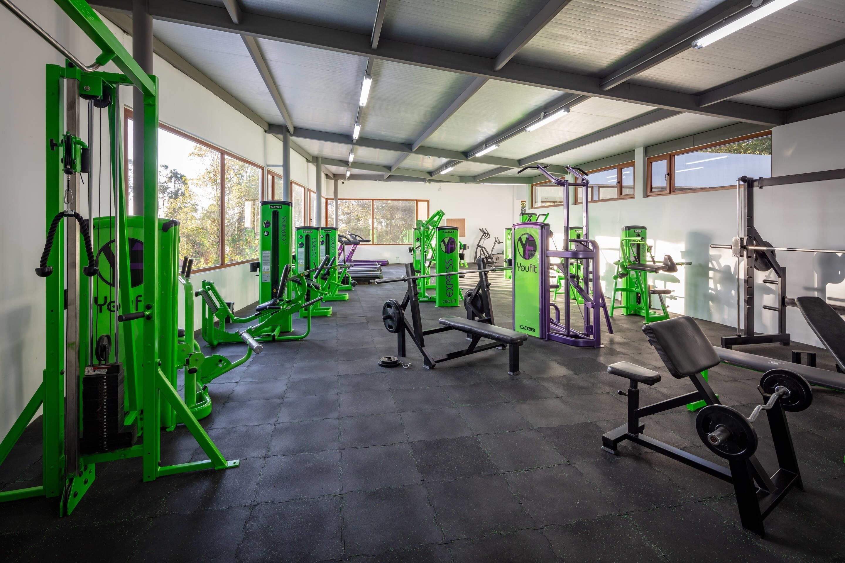 Un gimnasio con diversos equipos de ejercicio, como máquinas de pesas y bancos, dispuesto en un espacio amplio con ventanas grandes.