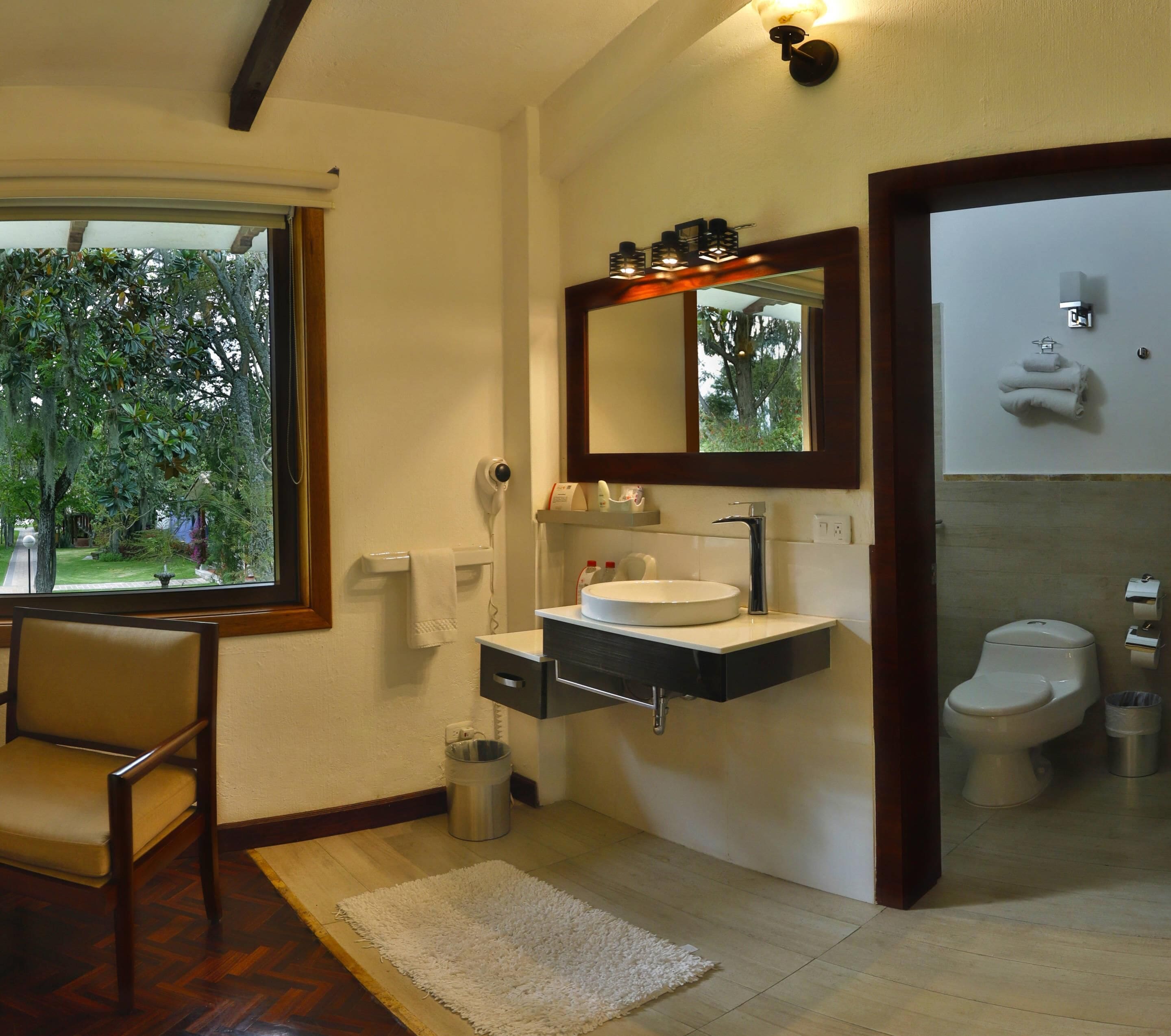 A modern bathroom with a large window, a sink with a mirror, a toilet, and a chair.